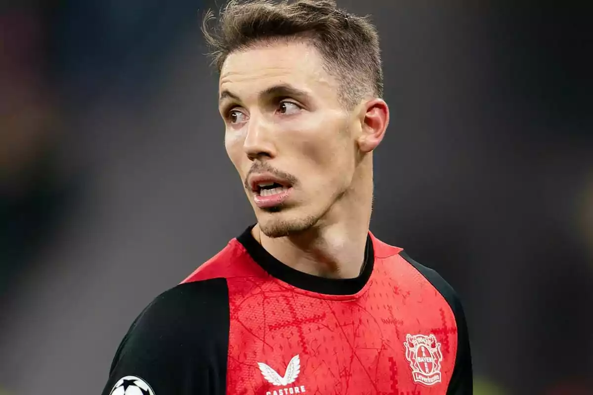 Un jugador de fútbol con uniforme rojo y negro del Bayer Leverkusen mira hacia un lado durante un partido. Un jugador de fútbol con uniforme rojo y negro del Bayer Leverkusen mira hacia un lado durante un partido.