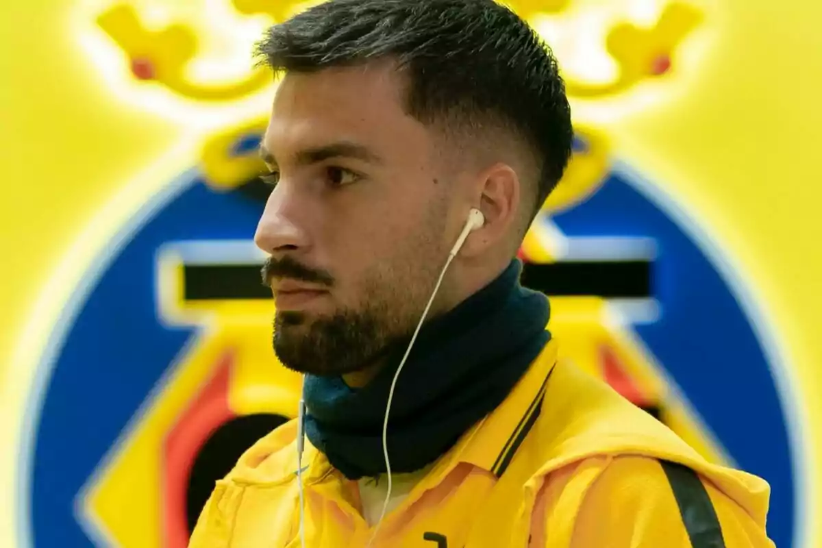 Un hombre con auriculares y bufanda, vestido de amarillo, frente a un fondo con un escudo deportivo.