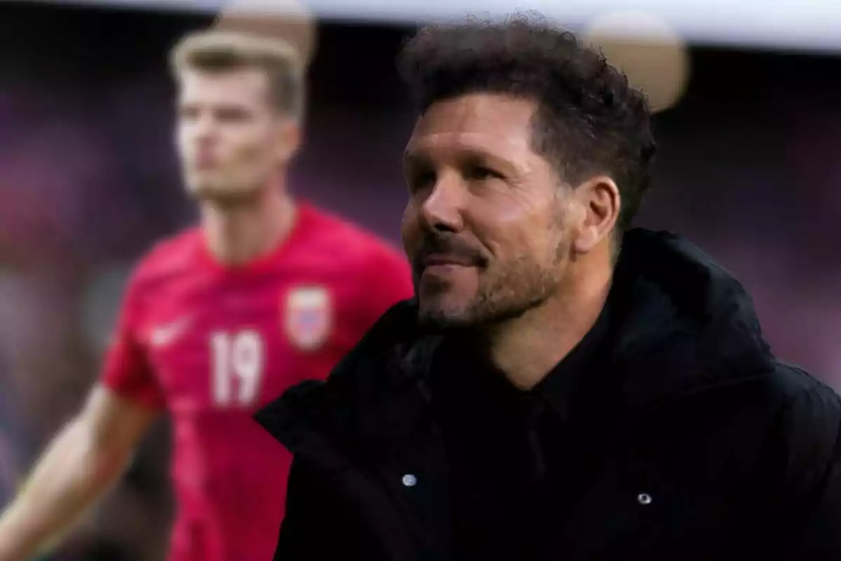 Un hombre con barba y cabello rizado, vestido con una chaqueta negra, está sonriendo mientras un jugador de fútbol con una camiseta roja y el número 19 en el pecho se ve desenfocado en el fondo.