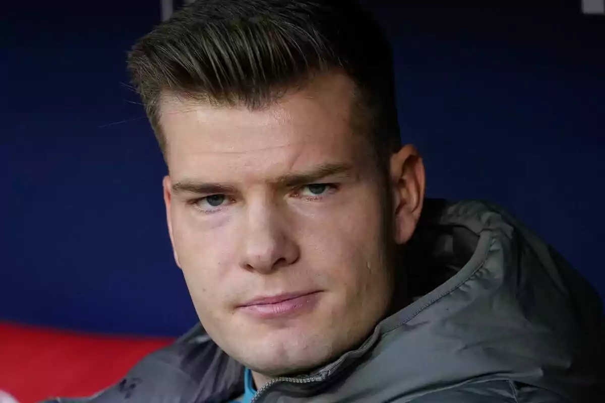 A man with a serious expression and short hair, wearing a gray jacket, against a dark background.