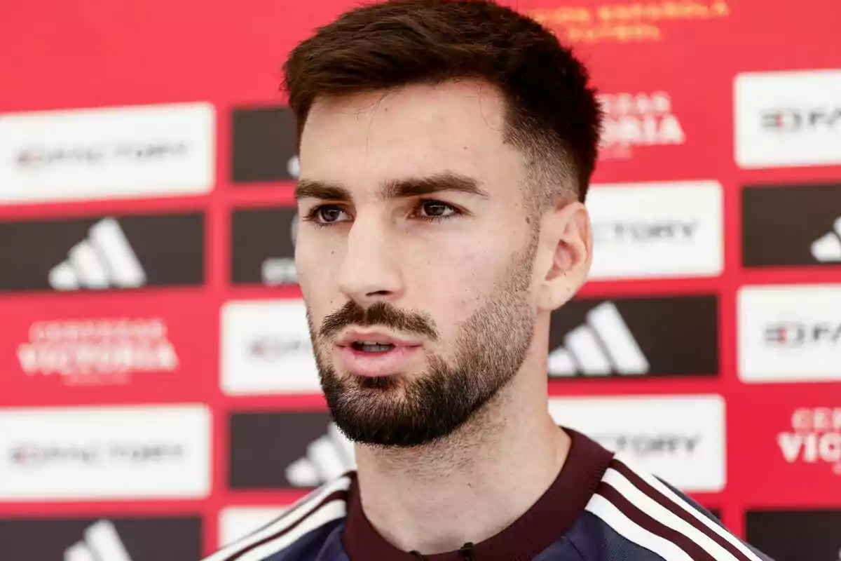 Un hombre con barba y cabello corto está hablando frente a un fondo rojo con logotipos.