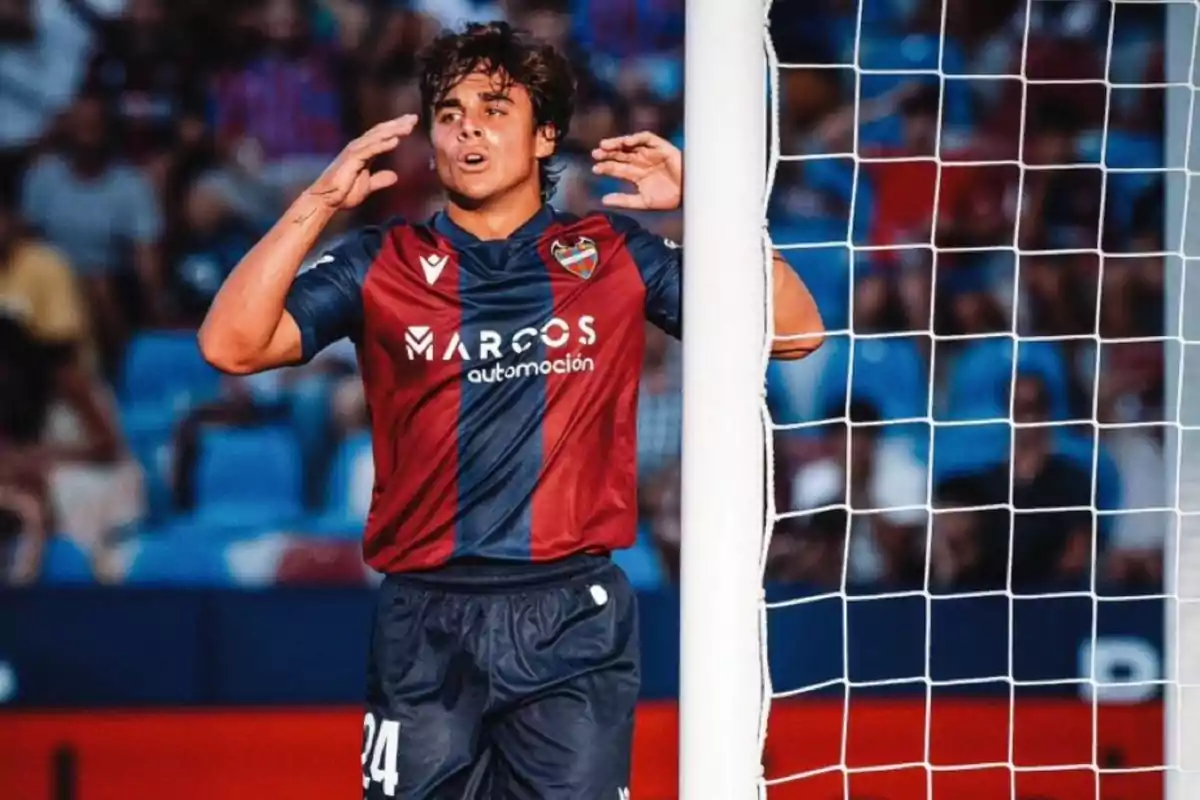 Un jugador de fútbol con uniforme azul y granate está junto a un poste de la portería en un estadio lleno de espectadores.