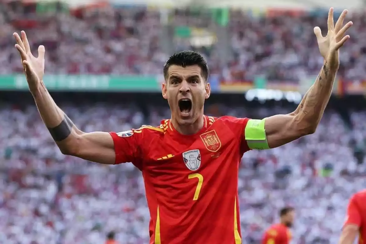 Jugador de fútbol con uniforme rojo celebrando con los brazos en alto en un estadio lleno de espectadores.