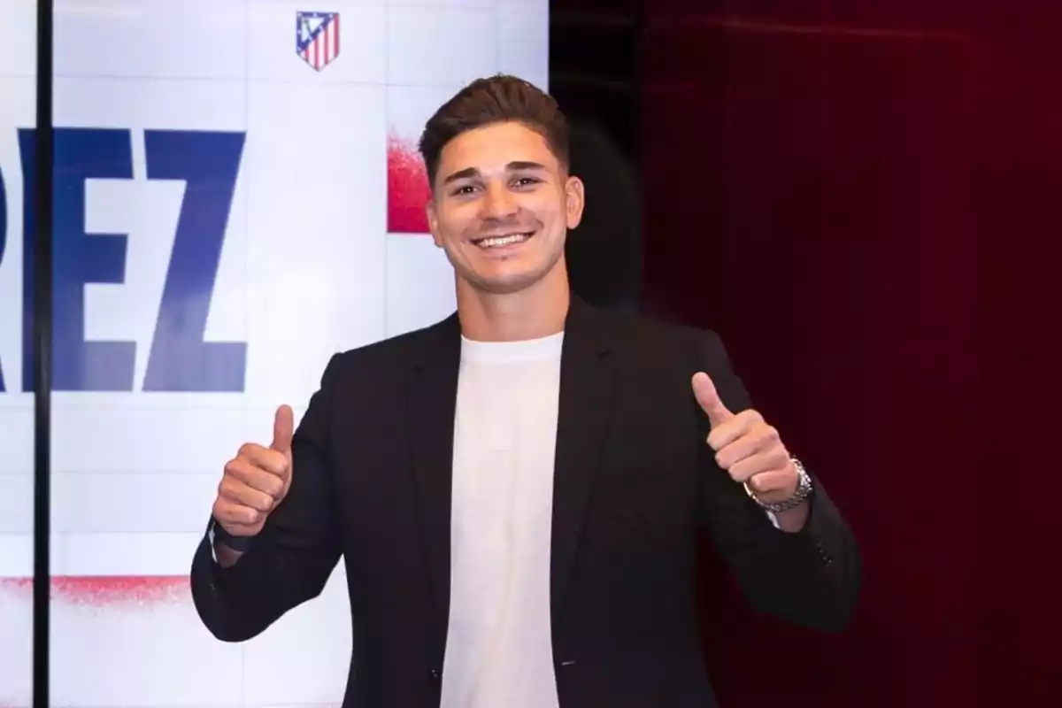 A smiling man in a black suit and white t-shirt, giving both thumbs up in front of a background with the Atlético de Madrid logo.