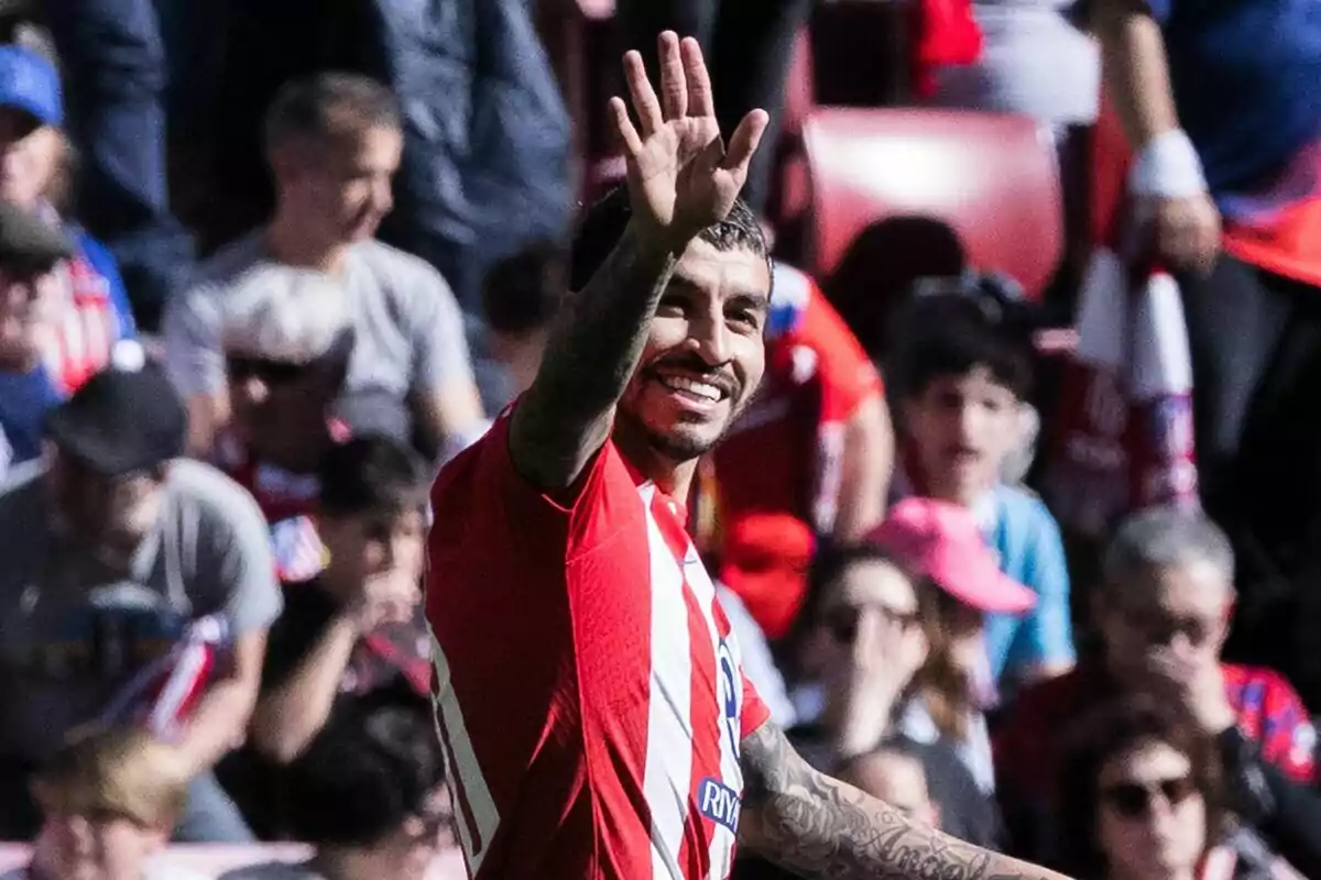 Un jugador de fútbol con la camiseta del Atlético de Madrid saluda sonriente a la multitud en un estadio lleno de aficionados.