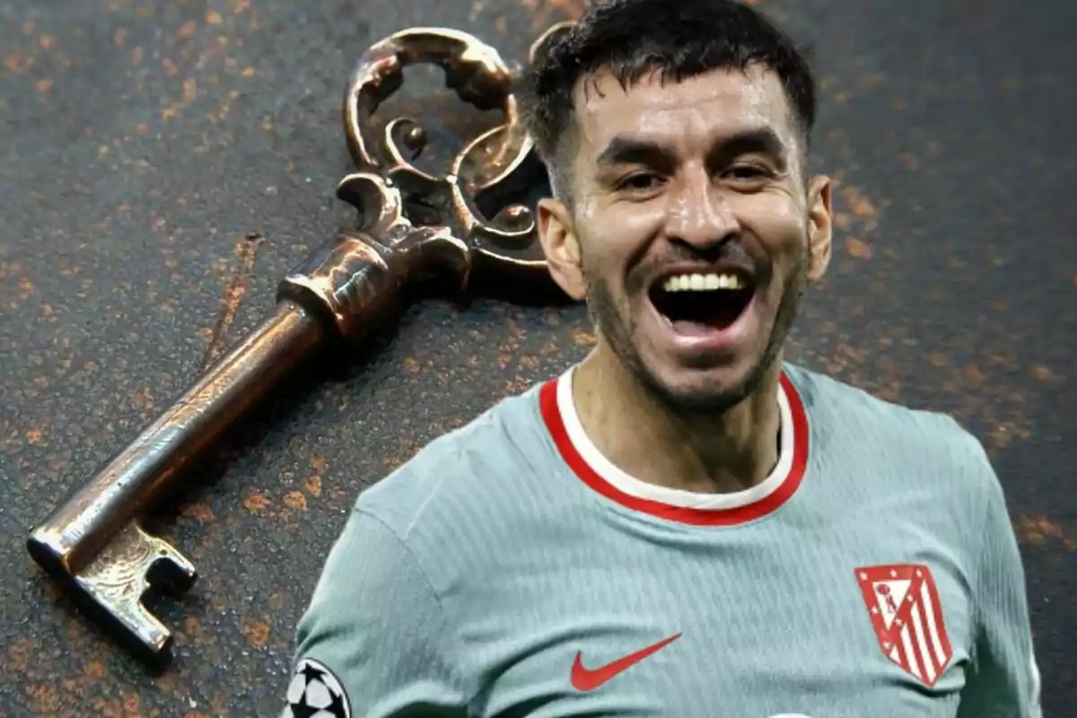 A smiling soccer player in a grey Atletico Madrid jersey stands next to an old key on a dark surface.