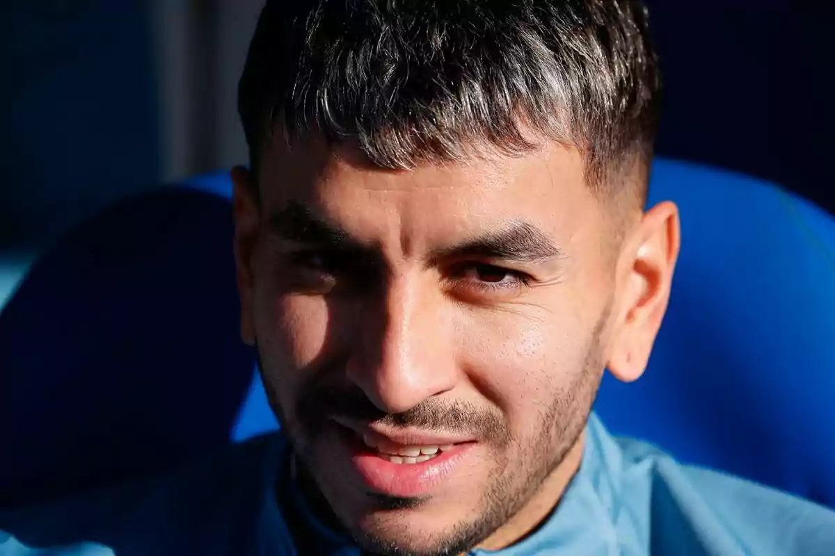Un hombre con cabello corto y barba ligera sonriendo mientras está sentado en un asiento azul.