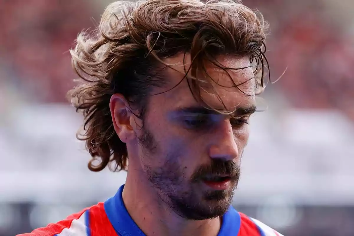 Un jugador de fútbol con cabello largo y barba, vistiendo una camiseta roja y azul, mira hacia abajo en un estadio.