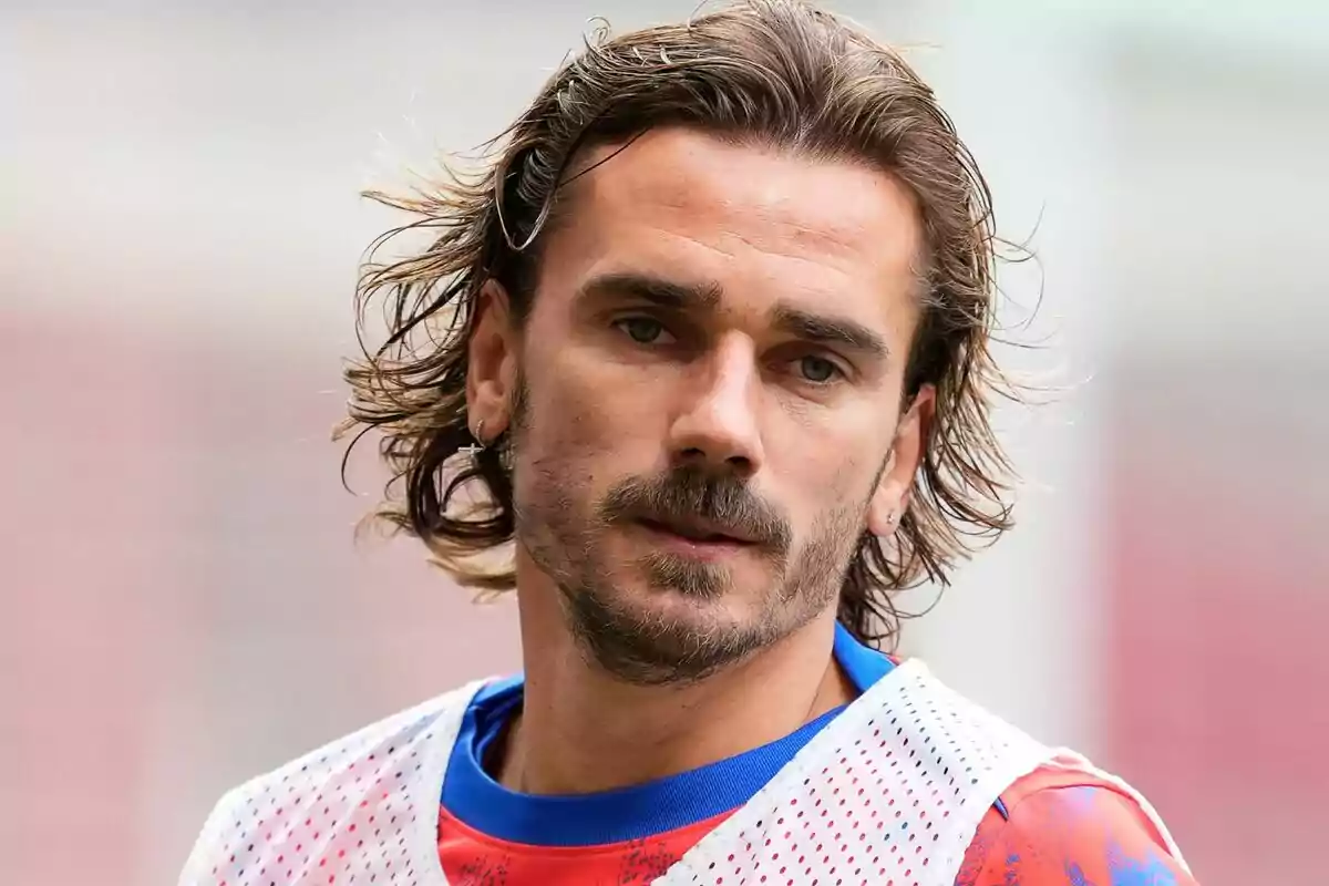 Un jugador de fútbol con cabello largo y barba viste una camiseta deportiva y un peto blanco.