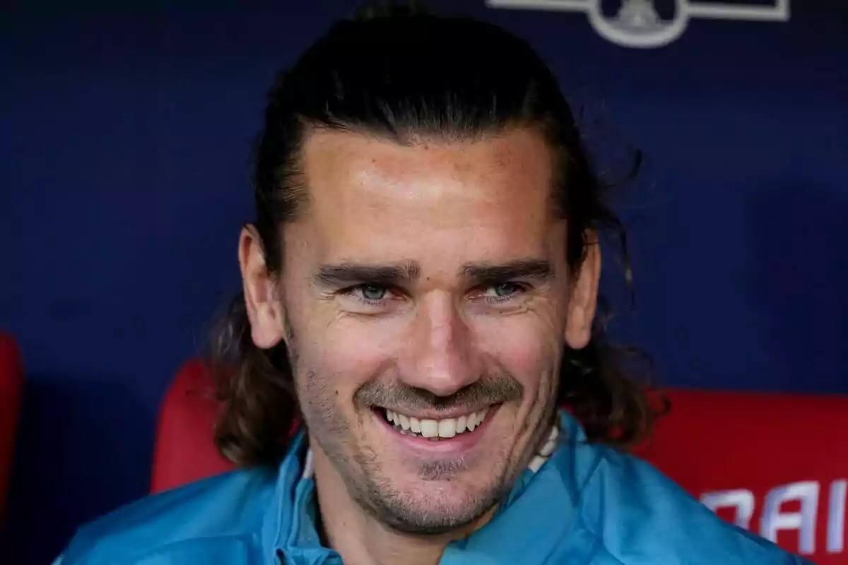 A man with long hair and a light beard smiles while sitting.