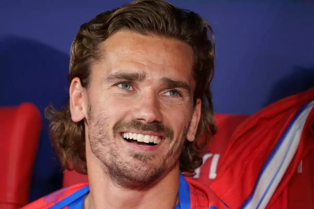 Hombre con barba y cabello largo sonriendo mientras está sentado en una silla roja.