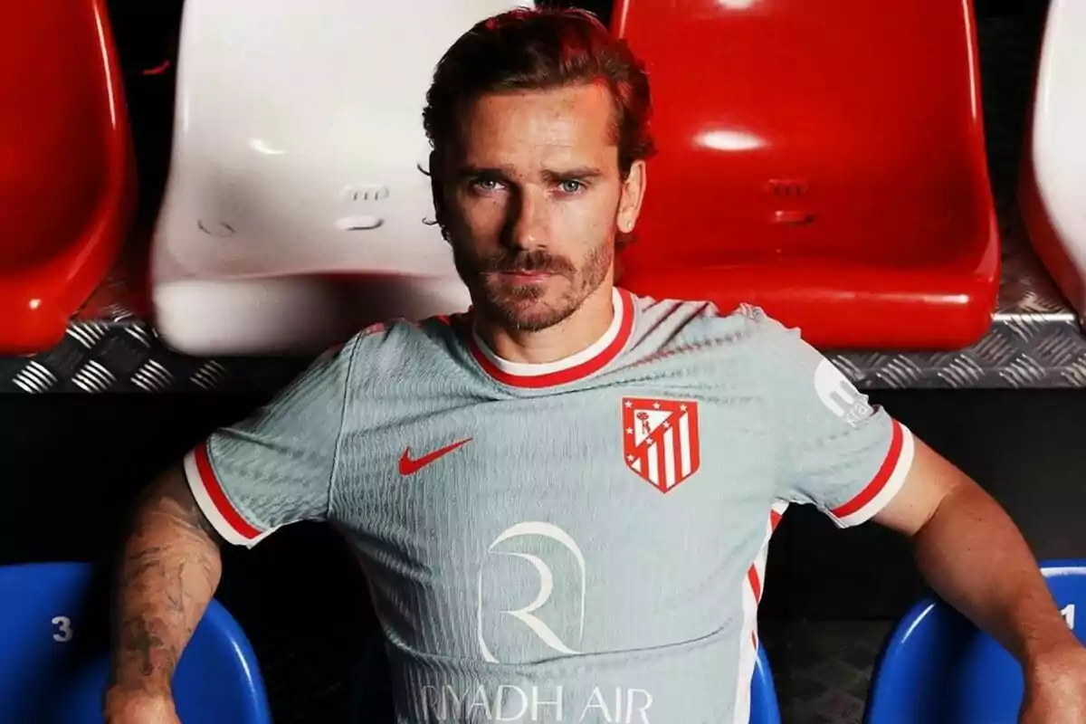 Un jugador de fútbol con la camiseta del Atlético de Madrid está sentado en una grada con asientos rojos, blancos y azules.