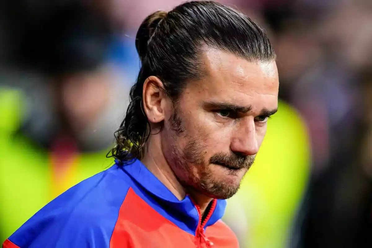 A man with long hair and a beard, wearing a blue and red jacket, appears focused as he looks ahead.