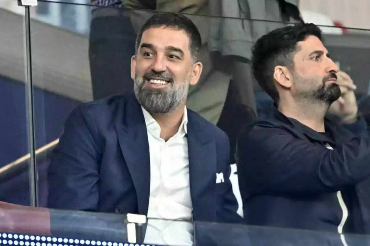 Two men sitting at a sporting event, one of them smiling while the other looks ahead.