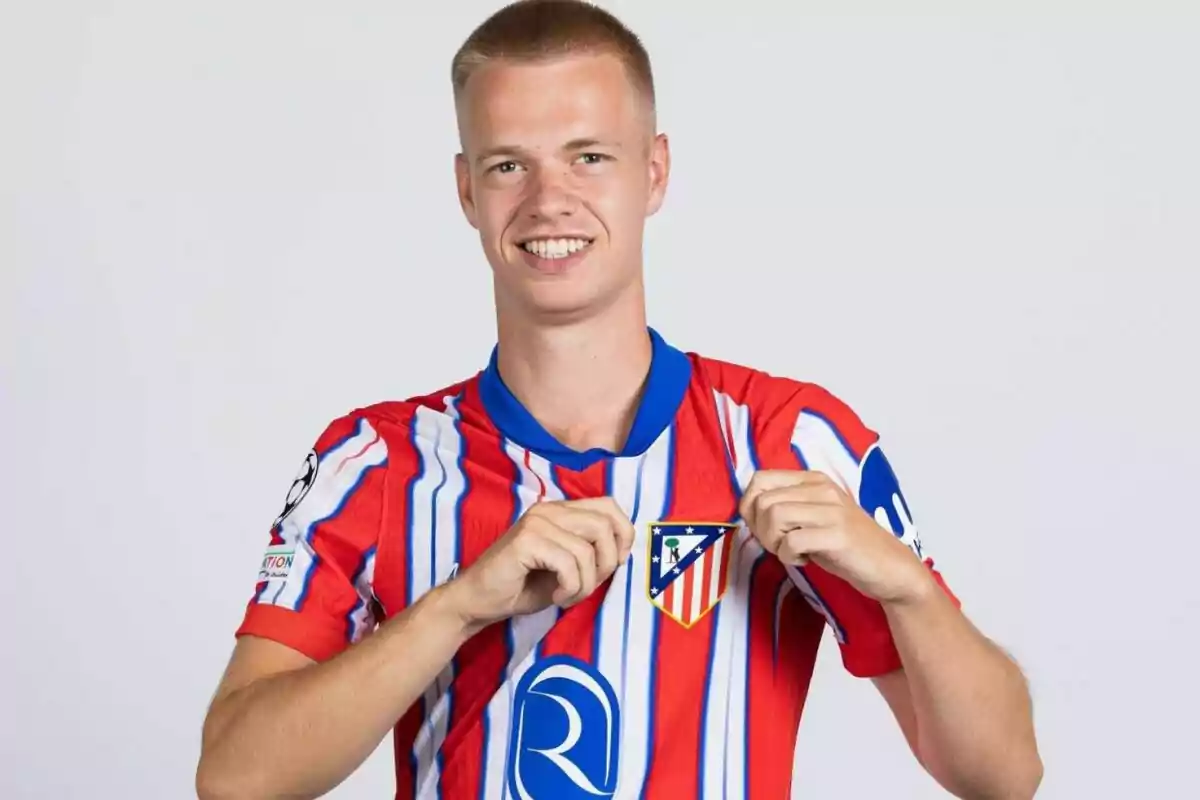 Un jugador de fútbol con una camiseta roja y blanca sostiene el escudo de su equipo mientras sonríe a la cámara.