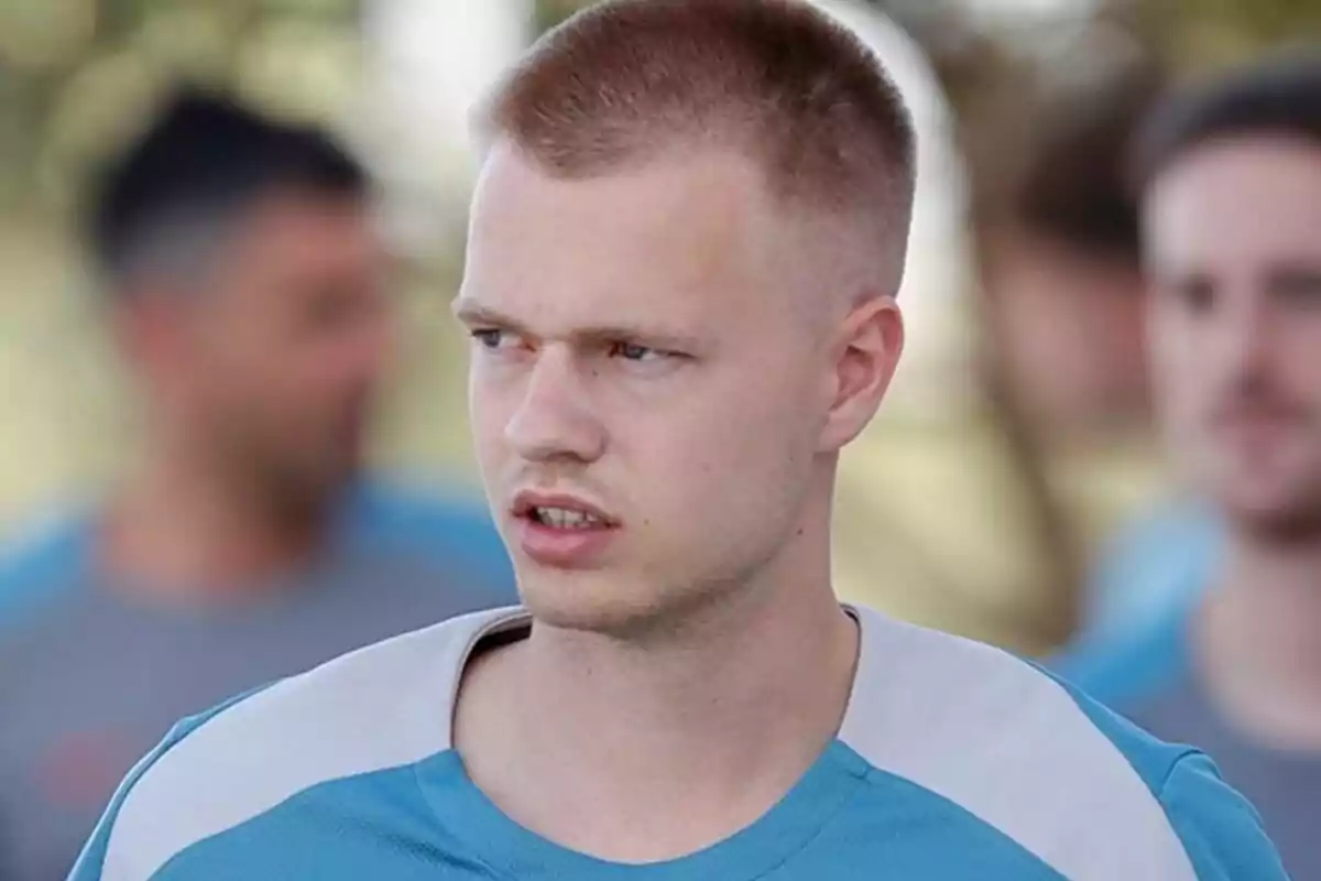 Un hombre con camiseta azul claro y corte de cabello corto, mirando hacia la derecha con expresión seria, con personas desenfocadas en el fondo.