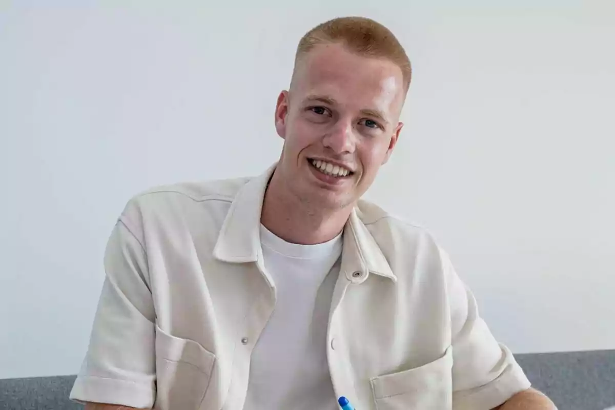 Un hombre joven con cabello corto y rubio, sonriendo y vistiendo una camisa beige sobre una camiseta blanca, sentado en un sofá gris con una pared blanca de fondo.