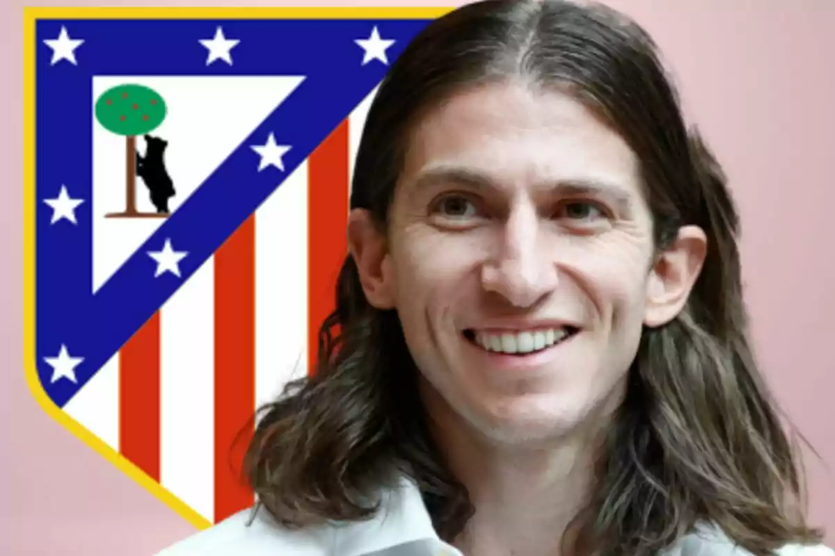 Un hombre sonriente con cabello largo y una camisa blanca está frente al escudo del Atlético de Madrid.