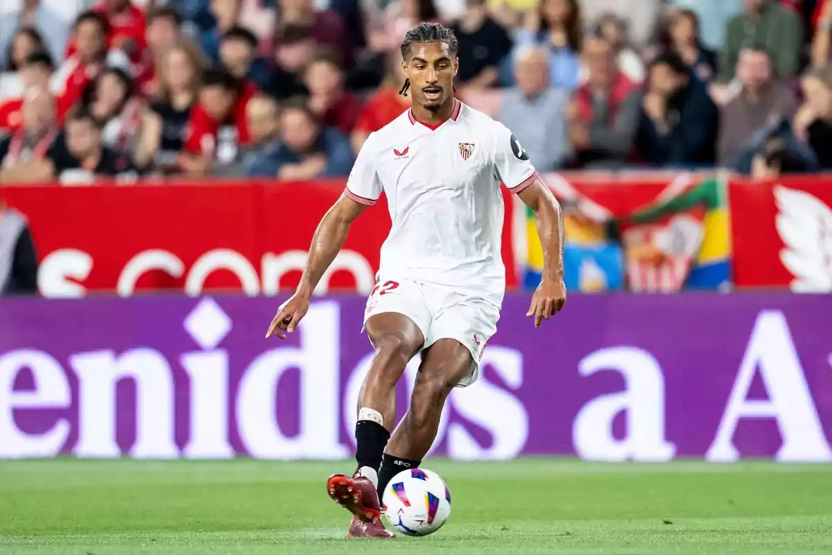 Un jugador de fútbol del Sevilla FC, vestido con el uniforme blanco del equipo, controla el balón en el campo durante un partido, con la multitud de espectadores en las gradas de fondo.