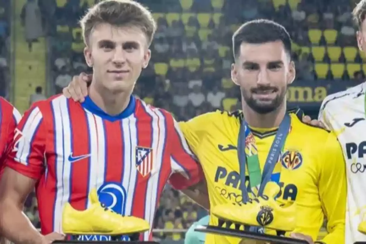 Dos jugadores de fútbol posan juntos con trofeos en forma de bota dorada, uno lleva la camiseta del Atlético de Madrid y el otro del Villarreal, en un estadio lleno de espectadores.