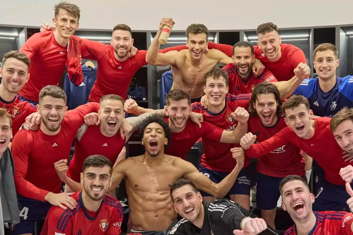 Un grupo de jugadores de fútbol celebrando en el vestuario, todos llevan camisetas rojas excepto uno que está sin camiseta, algunos están sonriendo y levantando los brazos en señal de victoria.