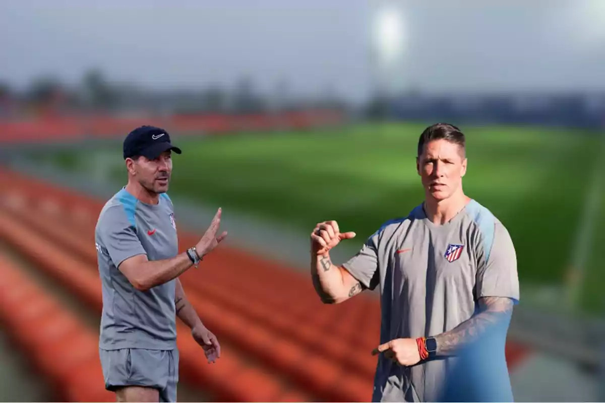 Dos hombres con ropa deportiva del Atlético de Madrid están en un campo de entrenamiento, uno de ellos lleva una gorra y el otro tiene tatuajes en el brazo.