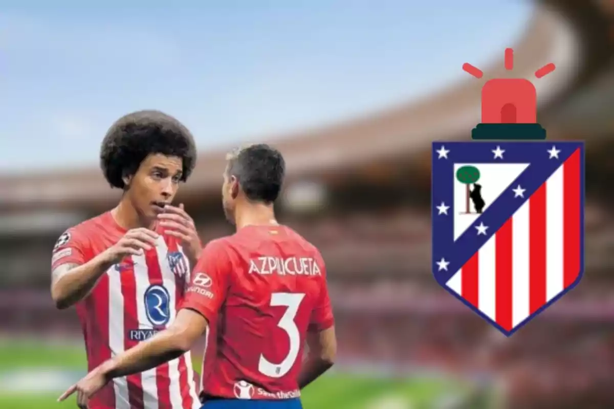 Dos jugadores del Atlético de Madrid conversando en el campo con el escudo del equipo a la derecha.