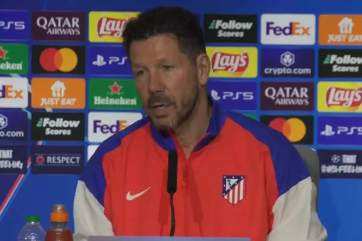 Un hombre con chaqueta del Atlético de Madrid en una rueda de prensa con logos de patrocinadores de fondo.