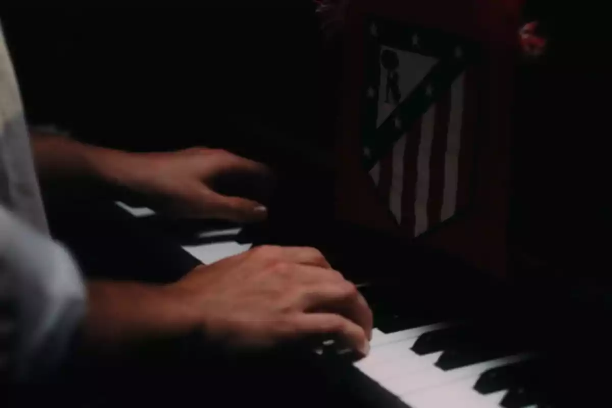 Manos tocando un piano con un escudo del Atlético de Madrid en el fondo.