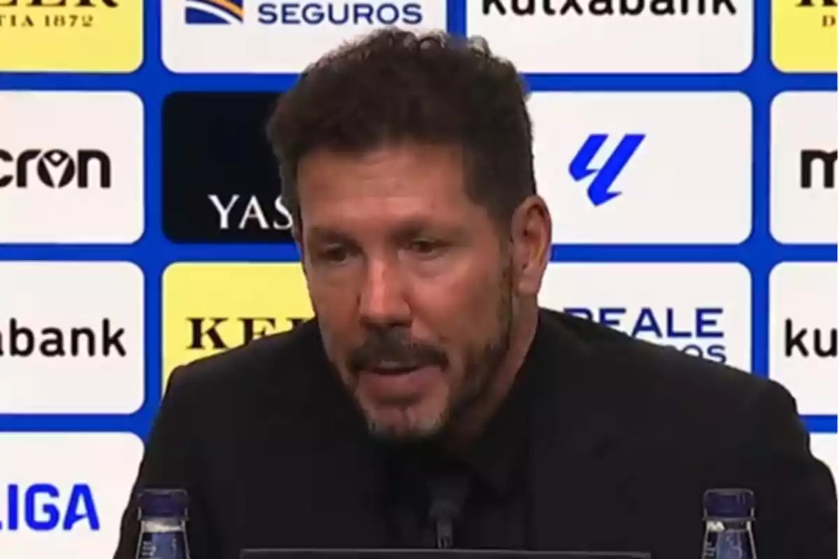 Un hombre con barba y cabello oscuro habla en una conferencia de prensa con un fondo de logotipos de patrocinadores.