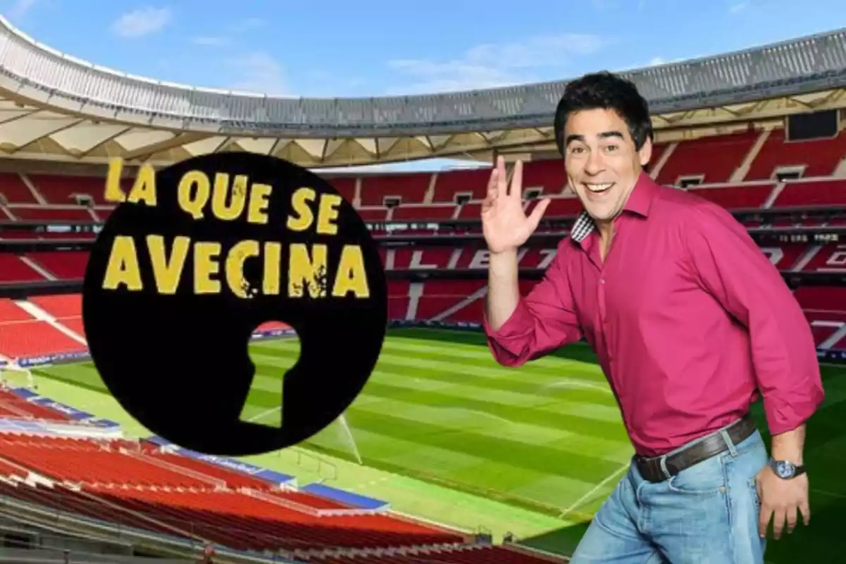 Un hombre sonriente con camisa rosa y jeans está posando en un estadio de fútbol con el logo de "La que se avecina" al lado.