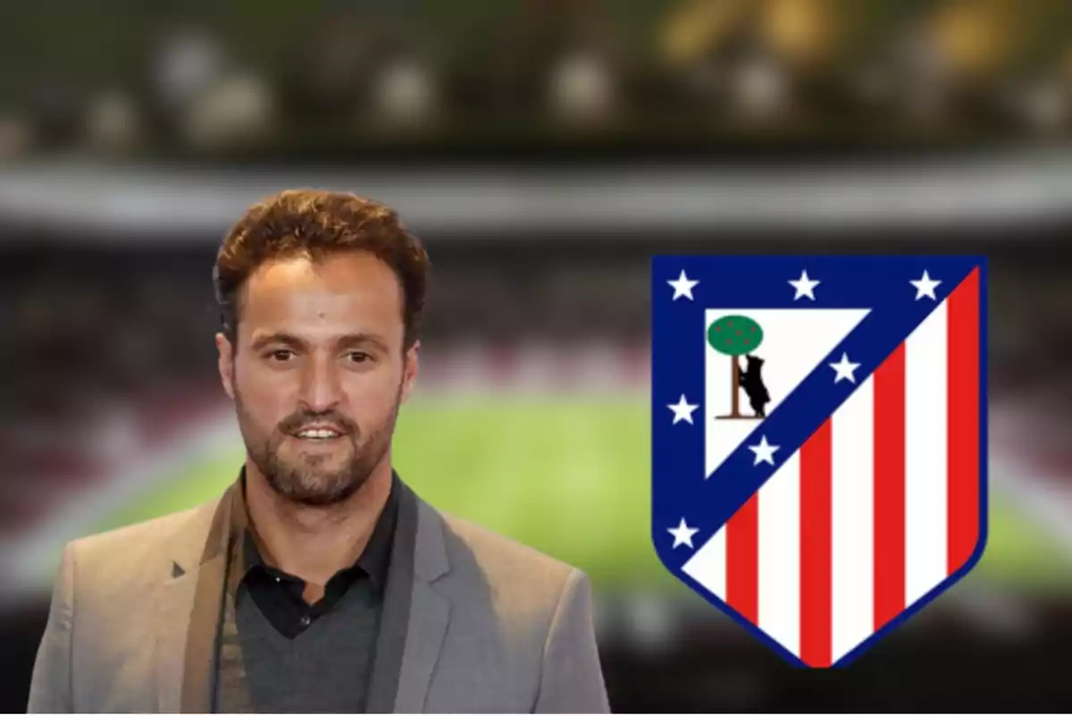 Un hombre con barba y cabello corto lleva un traje gris y está de pie junto al escudo del Atlético de Madrid en un estadio de fútbol.