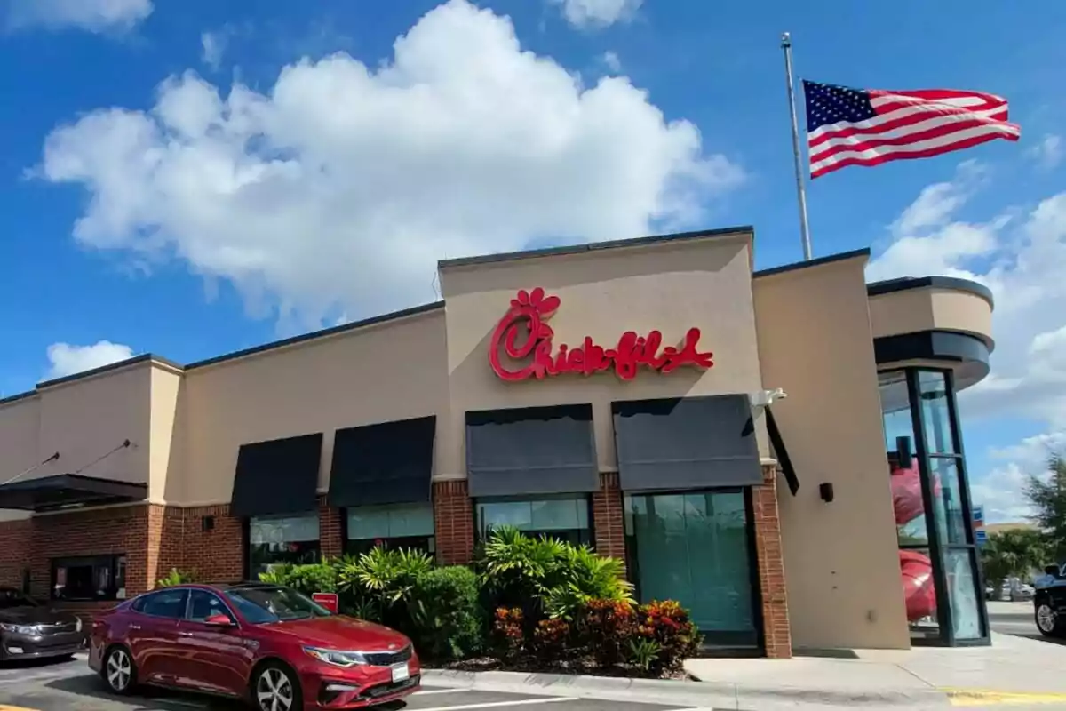 Un restaurante de comida rápida, que es Chick-fil-A, con un letrero rojo en la fachada y una bandera estadounidense ondeando en el cielo.
