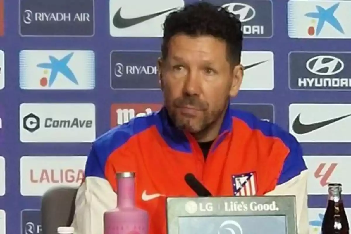 Un hombre con chaqueta del Atlético de Madrid en una rueda de prensa con logos de patrocinadores de fondo.