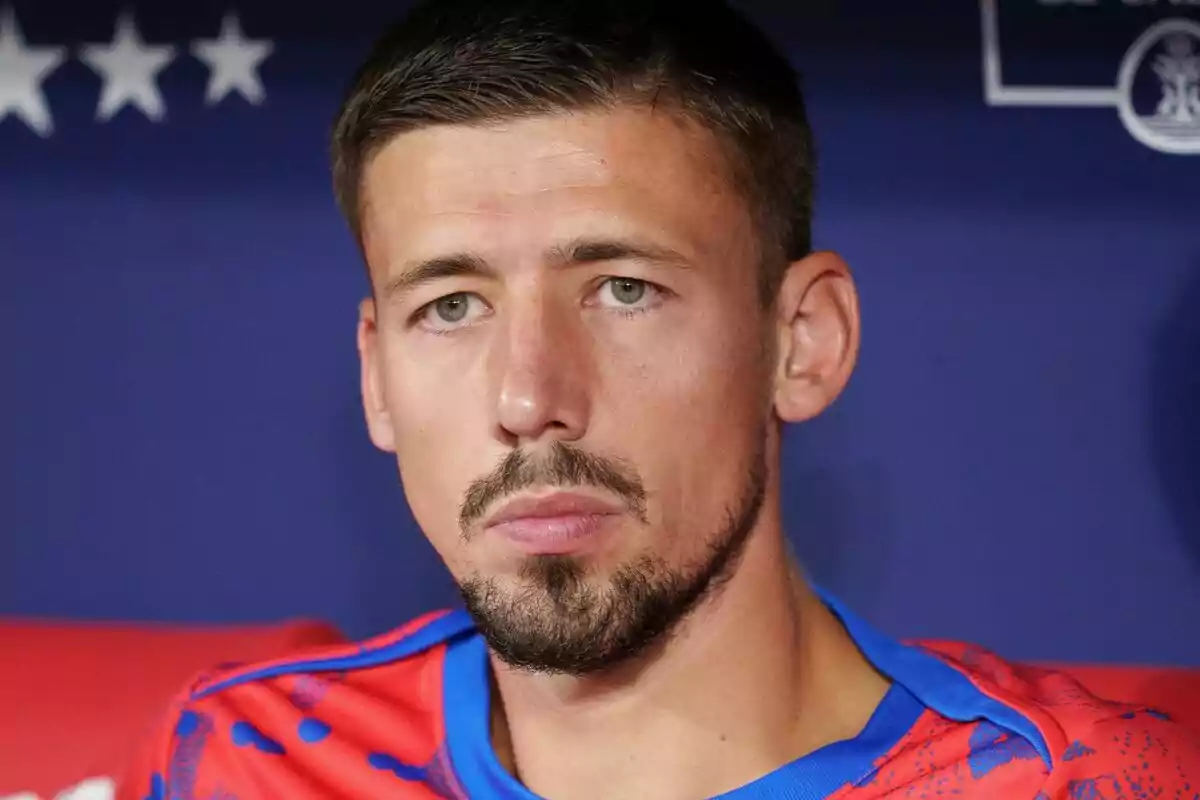 Un hombre con barba y cabello corto lleva una camiseta deportiva roja y azul frente a un fondo azul con estrellas blancas.