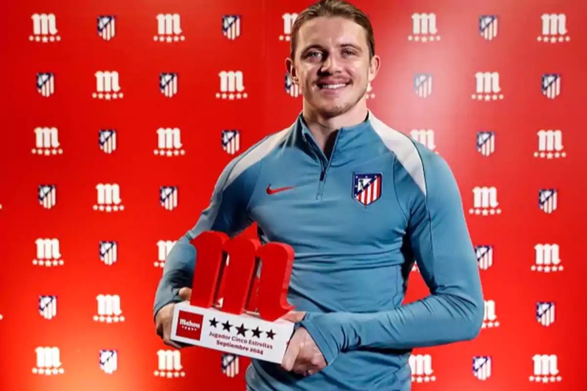 Un hombre con una sudadera del Atlético de Madrid sostiene un trofeo frente a un fondo rojo con logotipos del equipo y de Mahou.