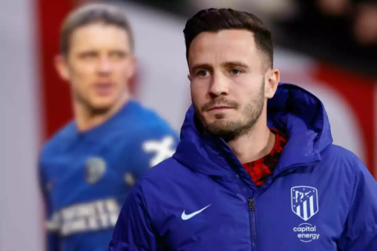 Un jugador de fútbol con una chaqueta azul del Atlético de Madrid aparece en primer plano, mientras que otro jugador con una camiseta azul se ve desenfocado en el fondo.