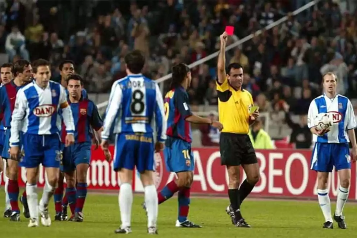 Un árbitro muestra una tarjeta roja a un jugador durante un partido de fútbol entre dos equipos con uniformes azulgrana y blanquiazul.