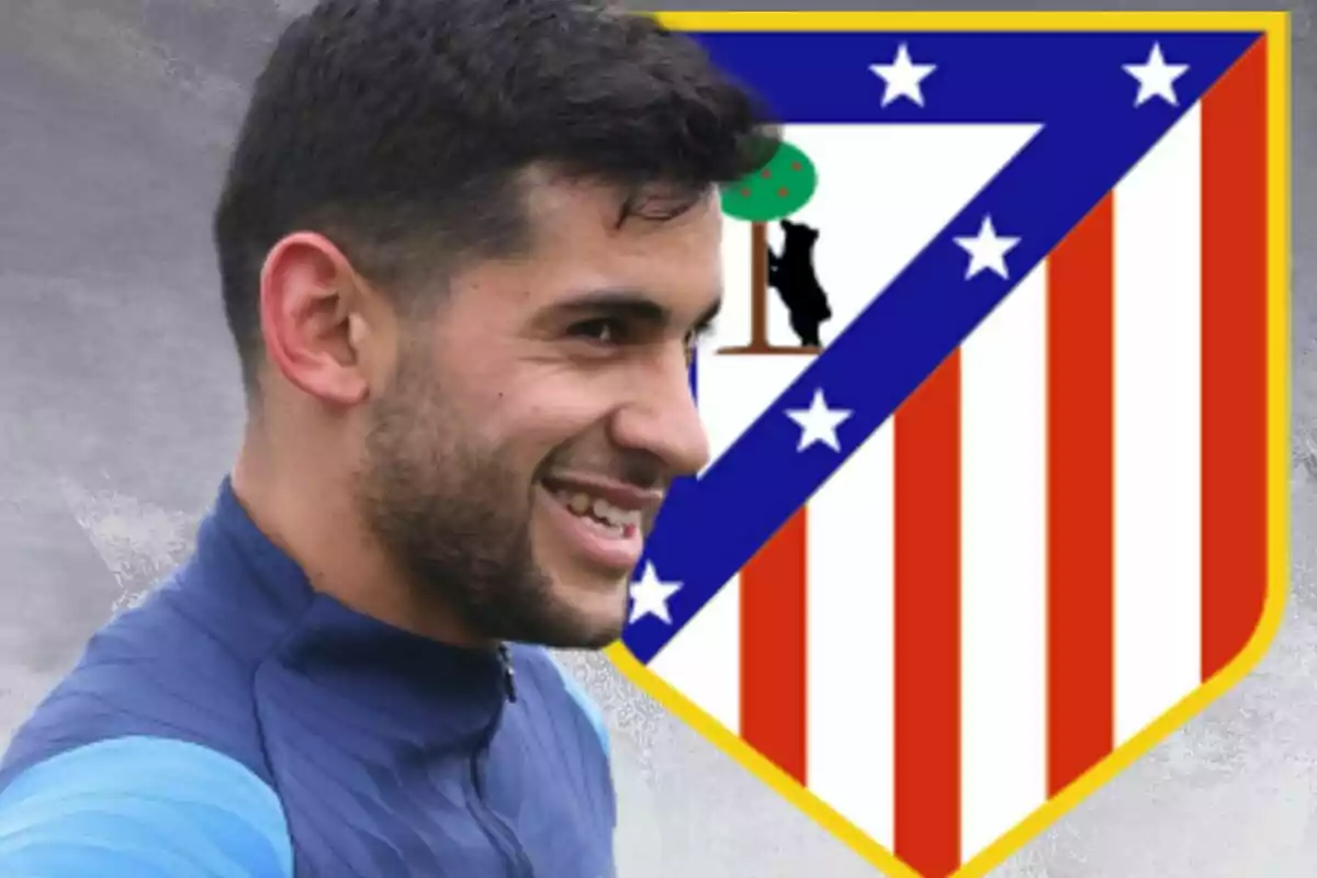 Un hombre sonriente con una chaqueta azul está frente a un escudo de un equipo de fútbol con rayas rojas y blancas y un oso junto a un árbol.