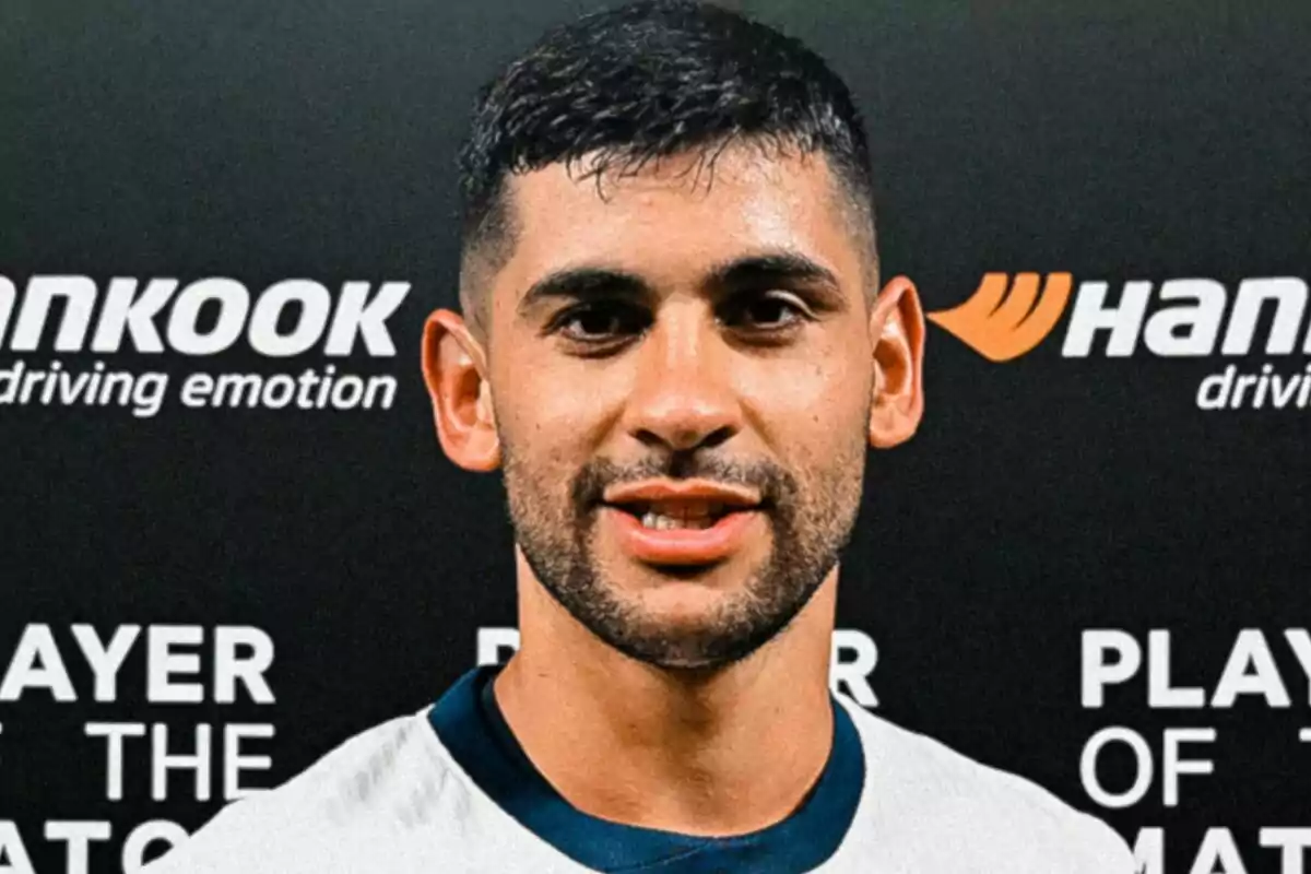 Un jugador de fútbol sonríe frente a un fondo con logotipos de patrocinadores.