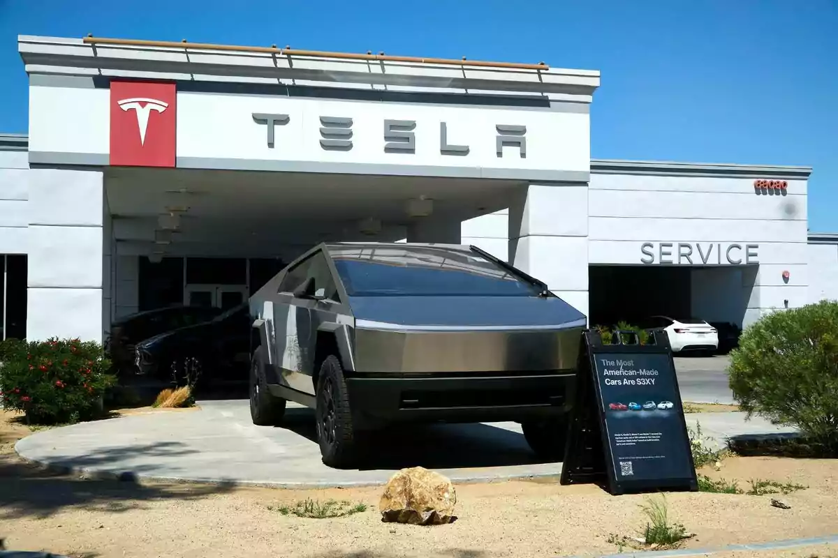 A Tesla Cybertruck pickup truck is parked in front of a Tesla dealership with a promotional sign next to it and the service building in the background. A Tesla Cybertruck pickup truck is parked in front of a Tesla dealership with a promotional sign next to it and the service building in the background.