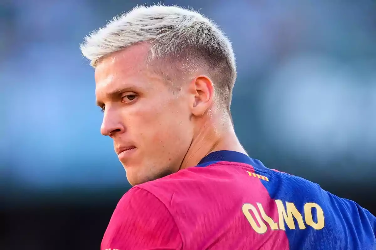 Un jugador de fútbol con cabello rubio y camiseta azul y roja mira hacia un lado. Un jugador de fútbol con cabello rubio y camiseta azul y roja mira hacia un lado.
