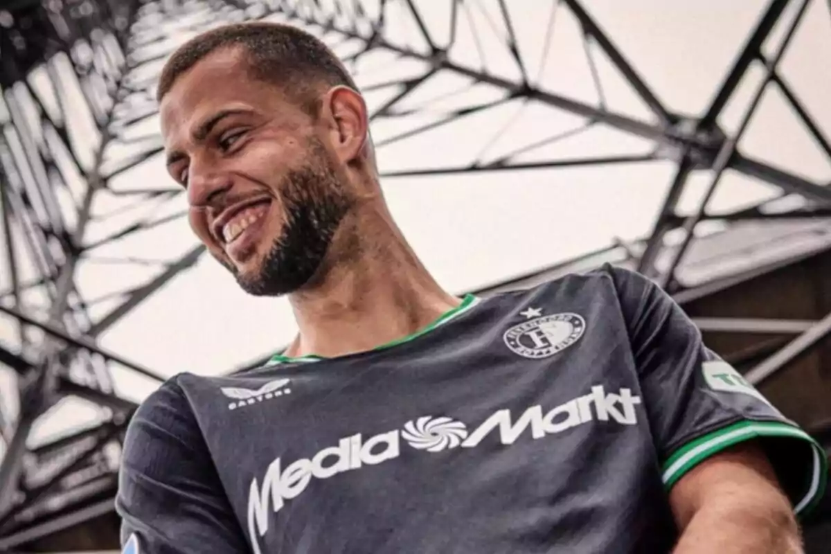 Un hombre con barba sonríe mientras viste una camiseta deportiva negra con el logo de MediaMarkt y un escudo en el pecho, con una estructura metálica en el fondo.