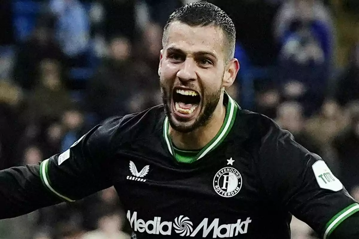 Jugador de fútbol celebrando un gol con expresión de euforia, vistiendo un uniforme negro con detalles verdes y el logo de un equipo en el pecho.