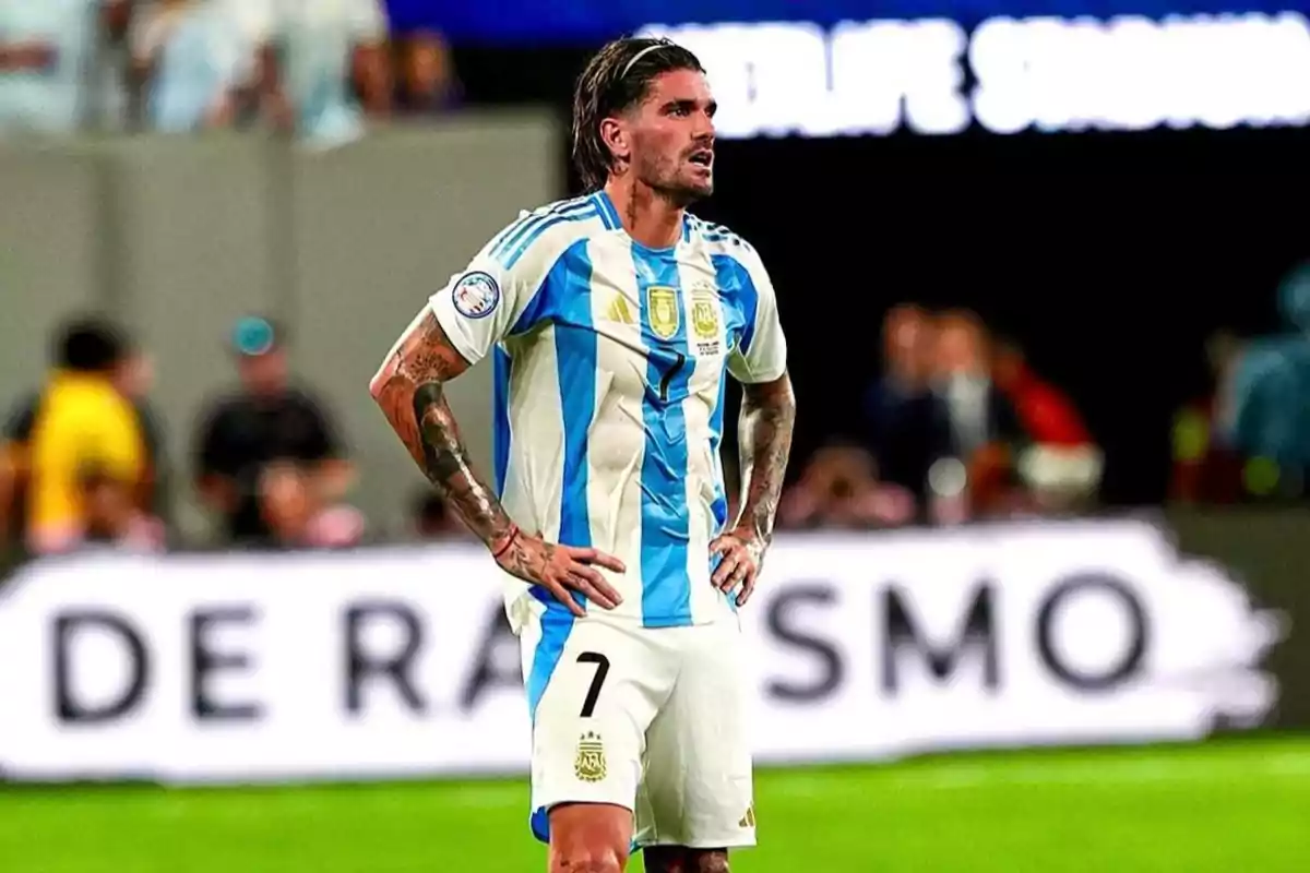 Un jugador de fútbol con el uniforme de la selección argentina está en el campo con las manos en la cintura y una expresión seria.