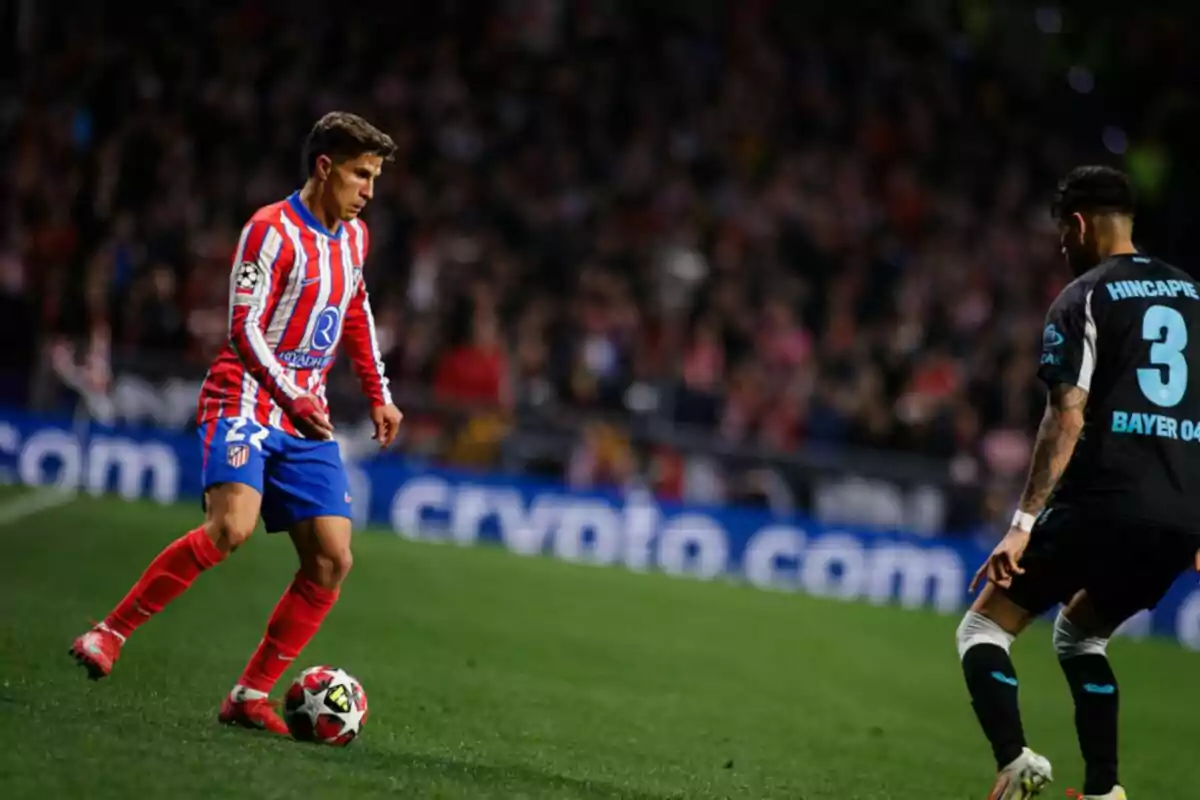 Dos jugadores de fútbol en acción durante un partido, uno con uniforme rojo y blanco y el otro con uniforme negro, en un estadio lleno de espectadores.