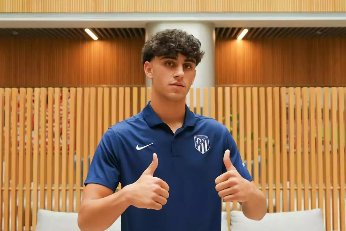 Un joven con una camiseta azul del Atlético de Madrid hace el gesto de pulgares arriba frente a una pared de listones de madera.