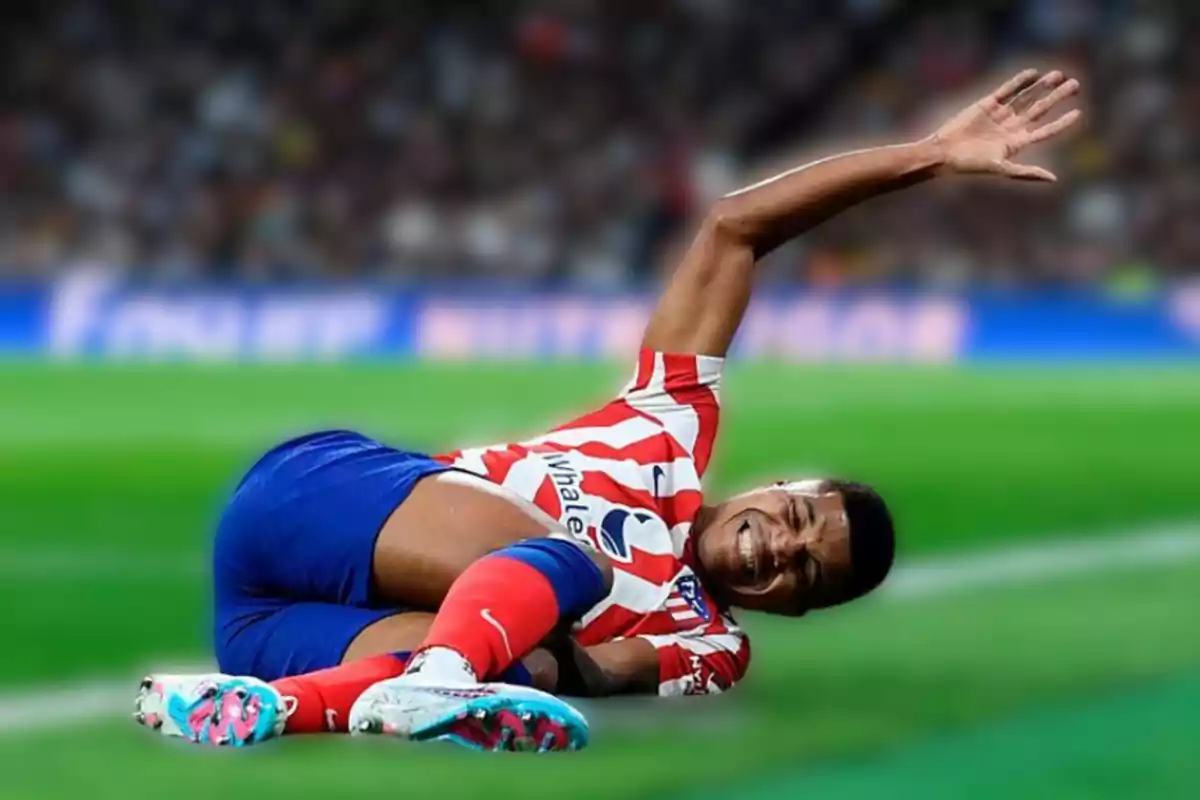 Un jugador de fútbol del Atlético de Madrid yace en el suelo con una expresión de dolor, levantando una mano mientras sostiene su pierna.