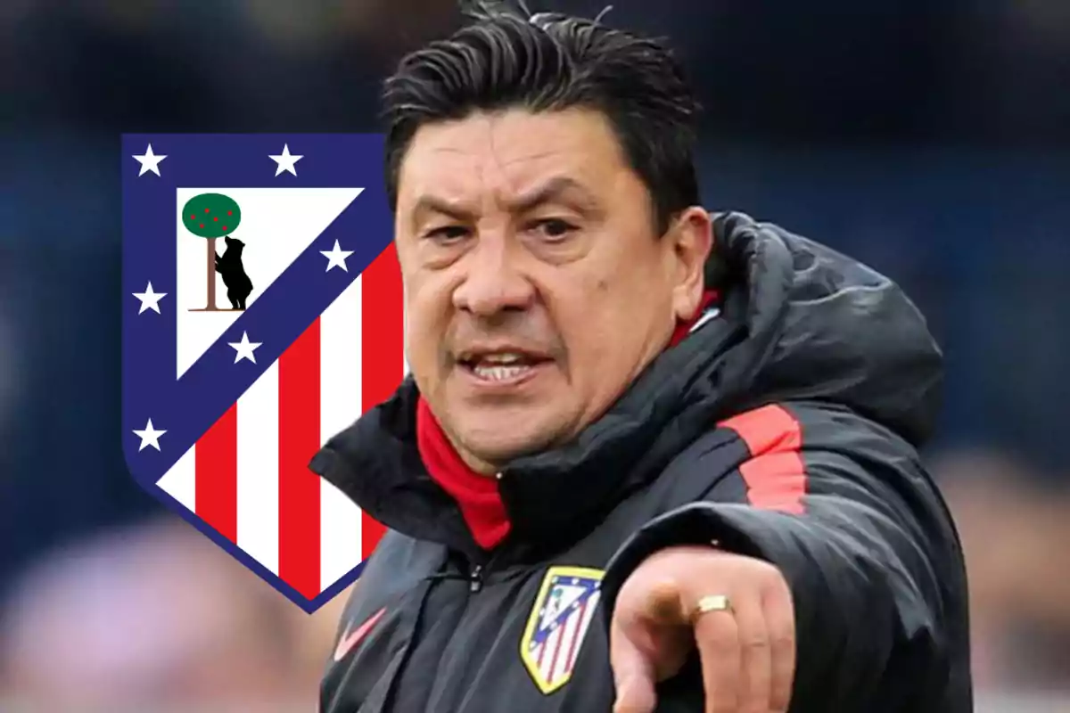 Un hombre con chaqueta deportiva negra y roja señalando hacia adelante con el escudo del Atlético de Madrid en el fondo.