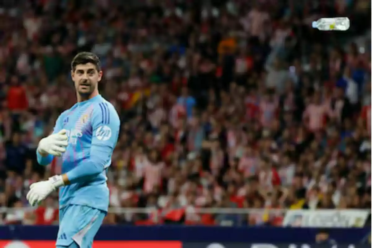 A soccer goalkeeper in a light blue uniform watches a plastic bottle in the air as the crowd in the stadium is blurred in the background.